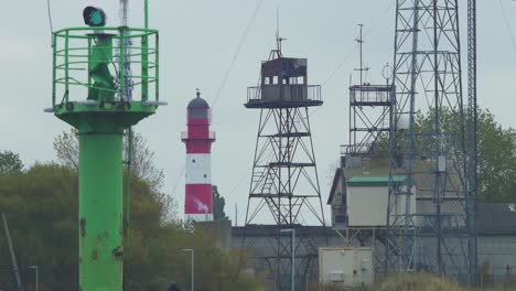 Red-painted-harbor-lighthouse-at-Port-of-Liepaja-on-a-overcast-day,-distant-view,-green-port-navigation-beacon-in-foreground,-communication-towers,-medium-shot