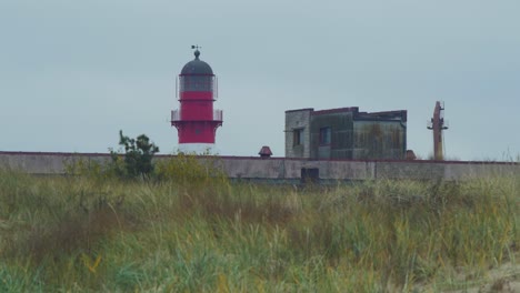 Red-painted-harbor-lighthouse-at-Port-of-Liepaja-on-a-overcast-day,-distant-view,-low-angle-medium-wide-shot-over-the-coastal-dunes-with-white-sand