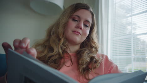 Caucasian-Woman-in-Pink-Sweater-Flips-Through-Pages-of-a-Book-on-Couch-by-Window-at-Daytime