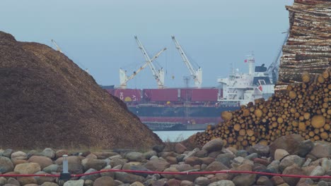 Large-cargo-vessel-loading-goods-at-Port-of-Liepaja-,-overcast-day,-piles-of-cut-wood-in-foreground,-distant-medium-shot