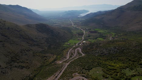 Hermoso-Valle-Con-La-Carretera-Entre-Campos-Verdes-Rodeados-De-Altas-Montañas-Y-El-Lago-Al-Fondo,-Albania