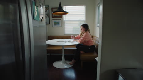 Stressed-out-Woman-Writing-at-Kitchen-Table-in-the-Afternoon-by-the-Window,-Barefoot-in-Pink-Sweater