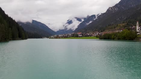 Flying-over-blue-lake-towards-small-town-surrounded-by-mountains-on-a-cloudy-day