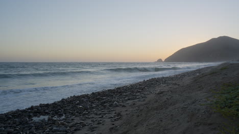 Stationary-shot-of-waves-rolling-through-the-Pacific-Ocean-with-the-sun-setting-behind-a-distant-mountain-located-in-Southern-California