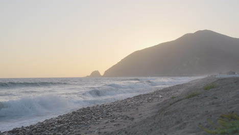 Stationary-slow-motion-shot-of-waves-crashing-onto-the-rocky-shore-of-Mondo's-Beach-at-sunset