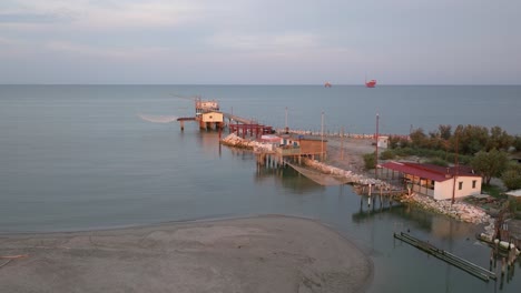 Slow-motion-aerial-view-of-fishing-huts-on-shores-of-estuary-at-sunset,italian-fishing-machine,-called-""trabucco"",Lido-di-Dante,-Ravenna-near-Comacchio-valley