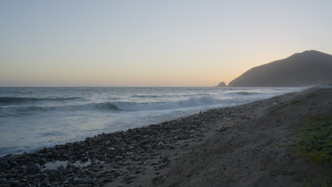 Stationärer-Schuss-Von-Wellen,-Die-In-Die-Felsen-Von-Mondos-Strand-Schlagen,-Mit-Sonnenuntergang-Hinter-Einem-Berg-Im-Hintergrund-In-Südkalifornien