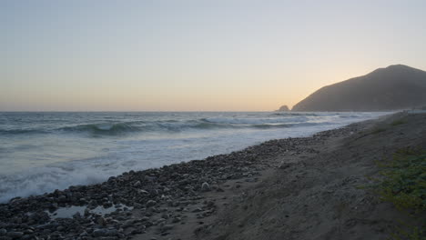 Stationary-shot-of-waves-crashing-onto-the-shores-of-Mondo's-Beach-with-the-sun-setting-behind-a-mountain-in-the-background-located-in-Southern-California