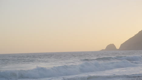 slow-motion-panning-shot-of-waves-rolling-in-along-Mondo's-Beach-as-the-sun-sets-behind-a-mountain-at-golden-hour-located-in-southern-California