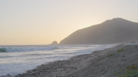 Stationary-slow-motion-shot-of-waves-running-along-the-shores-of-Mondo's-Beach-as-the-sun-sets-behind-mountain-located-in-Southern-California