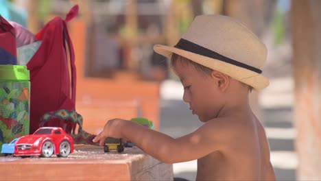Young-latin-baby-boy-wearing-a-hat-playing-with-his-car-toys-on-a-bench-at-the-beach