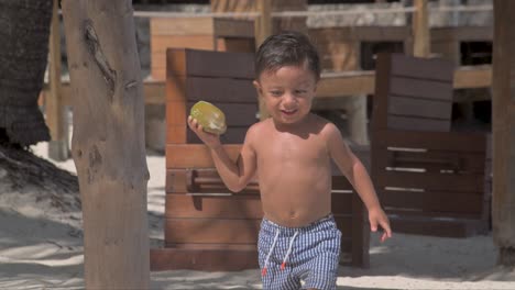 Junger-Lateinischer-Junge,-Der-An-Einem-Sonnigen-Tag-Am-Strand-Spazieren-Geht,-Seine-Badeanzug-Shorts-Trägt-Und-Eine-Kokosnuss-Trägt