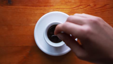 Abwärtsschuss-Einer-Hand,-Die-Zucker-In-Einer-Heißen-Espressotasse-Rührt,-Die-Auf-Einem-Braunen-Holztisch-Steht