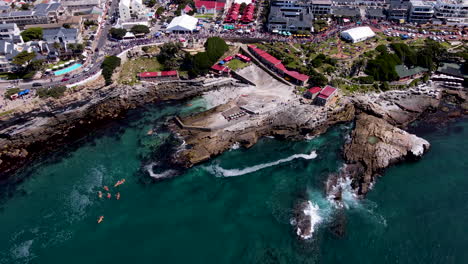 Kayaks-depart-rocky-shoreline-with-drone-view-of-Hermanus-Whale-Festival-parade