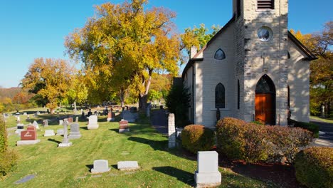 Old-Small-Catholic-Church-With-Cemetry-Headstones-and-Bright-Autumn-Trees