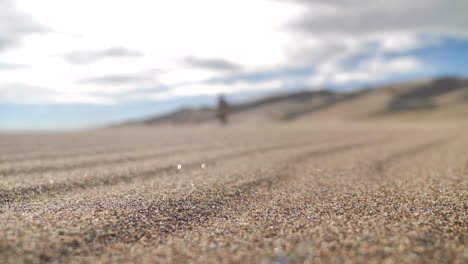 Close-Up-of-Sand-Blowing-with-Blurred-Figure-Walking-Towards-Camera-in-Desert