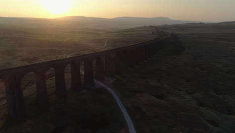 Aerial-Drone-Shot-into-Sun-of-Ribblehead-Viaduct-Train-Bridge-at-Stunning-Sunrise-in-Summer-in-Yorkshire-Dales-England-UK-with-Hills-in-Background