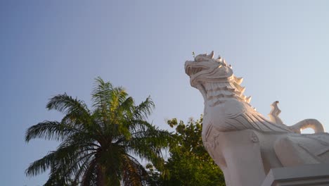 Looking-up-at-white-dragon-lion-statue-against-blue-sky-with-palm-tree