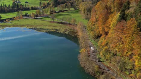 Aerial-View-Over-Autumnal-Forest-Trees-Beside-Lake-And-Road-Surrounding-It