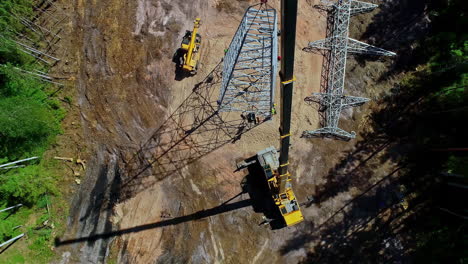 Aerial-drone-view-revealing-crane-placing-high-voltage-tower-in-vertical-position-in-forest-environment