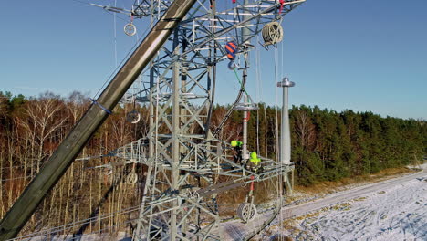 Aerial-dolly-out-revealing-two-electricians-working-in-the-maintenance-of-high-voltage-tower-in-snowy-forest