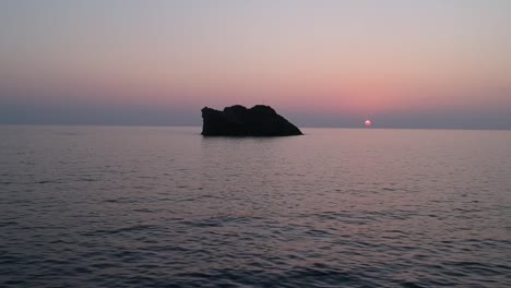 Ein-Einsamer-Felsen-Mitten-Im-Ozean-Bei-Sonnenuntergang,-Mit-Ruhigen,-Plätschernden-Wellen-Und-Einem-Farbenfrohen-Himmel