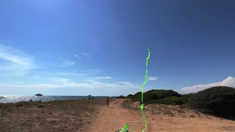 Little-girl-on-summer-holidays-playing-with-green-toy-kite-with-long-tail-in-blue-sky-holding-handles-and-sitting-on-small-chair-on-sandy-beach