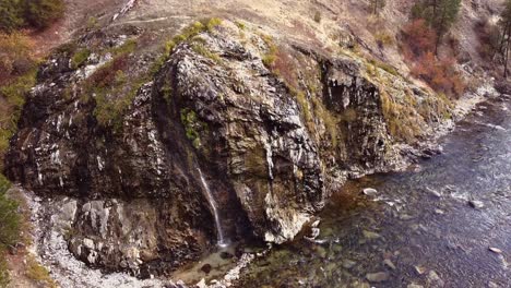 Aerial-drone-shot-approaching-a-hot-spring-waterfall-panning-down-rising-above-in-Boise-National-Forest-in-Idaho-with-the-spring-flowing-into-a-pool-below-beside-boise-river