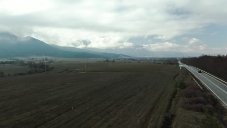 Drone-Disparó-Sobre-La-Carretera-Rural-Con-Coches-En-Movimiento-Y-Vistas-A-Las-Montañas-Con-Cielo-Nublado