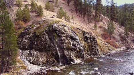 Aerial-drone-shot-approaching-a-hot-spring-waterfall-in-Boise-National-Forest-in-Idaho-located-on-Boise-river-with-steam-rising-and-flowing-water-below