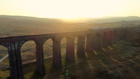 Luftdrohnenaufnahme-Des-Zuges,-Der-Auf-Der-Ribblehead-Viadukt-Zugbrücke-Bei-Atemberaubendem-Sonnenaufgang-Im-Sommer-In-Den-Yorkshire-Dales,-England,-Großbritannien,-Mit-Hügeln-Im-Hintergrund-Unterwegs-Ist