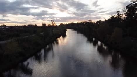 Drone-flying-over-river-during-sunset-near-a-forrest