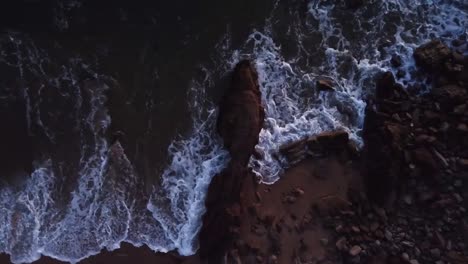 Top-view,-drone-hoovering-a-rock-at-the-beach,-water-hitting-rock-during-sunset