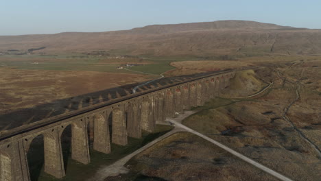 Aerial-Drone-Shot-Pulling-Back-from-Ribblehead-Viaduct-Train-Bridge-at-Stunning-Sunrise-with-Long-Shadows-in-Summer-in-Yorkshire-Dales-England-UK-with-3-Peaks-Whernside-Mountain-in-Background