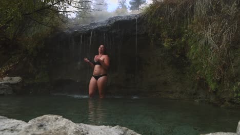 Woman-stands-beneath-a-thermal-hot-spring-waterfall-enjoying-the-benefits-of-natural-heated-hot-spring-water-on-body-in-a-heated-pool-in-nature-in-pacific-northwest