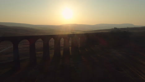 Luftdrohnenaufnahme,-Die-Sich-Von-Der-Ribblehead-Viadukt-Zugbrücke-Bei-Atemberaubendem-Sonnenaufgang-Im-Sommer-In-Den-Yorkshire-Dales,-England,-Großbritannien,-Mit-Hügeln-Im-Hintergrund-Zurückzieht