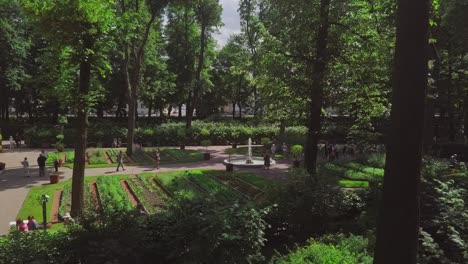 Summer-Garden,-an-aerial-drone-footage-of-a-fountain-through-trees-and-garden,-people-walking-in-a-park,-sun-flares,-someone-is-taking-a-selfie,-vegetables-growing