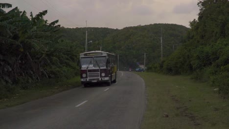 A-bus-driving-down-a-small-country-road-in-Cuba-surrounded-by-mountainous-forests