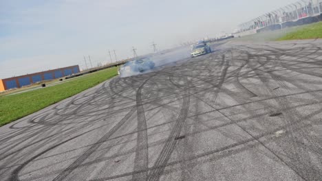 Two-green-muscle-drifting-cars-on-a-race-track-doing-a-parallel-drift-around-each-other-with-deep-tire-marks-on-asphalt,-slow-motion