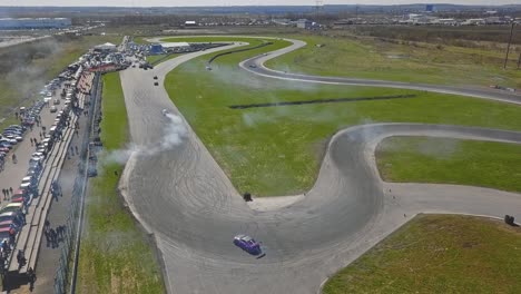 Race-cars-going-in-a-drift-over-a-turn-with-smoke-from-tires-and-viewers-sunny-weather,-wide-shot-of-a-map