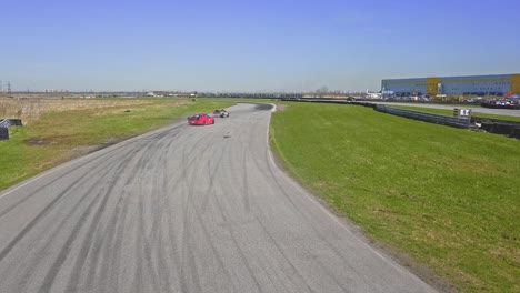 Aerial-drift-cars-passing-by-on-a-race-track,-red-and-brown-car-with-tire-skids-and-green-grass