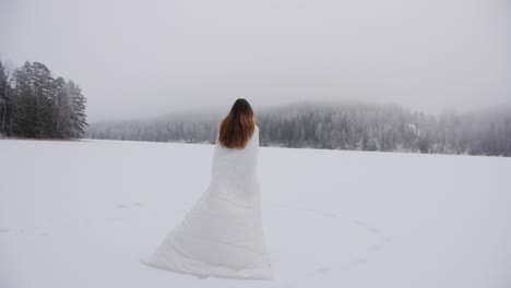 Woman-in-white-snowy-outfit-walking-over-a-frozen-lake-in-slow-motion-with-a-mist-over-forest-in-the-background