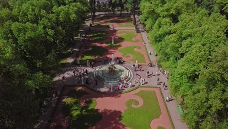 Summer-Garden,-an-aerial-drone-fly-by-of-a-beautiful-Koronniy-fountain-from-top,-people-visiting-the-festival,-with-flowers-and-summer-feeling,-river-in-the-background