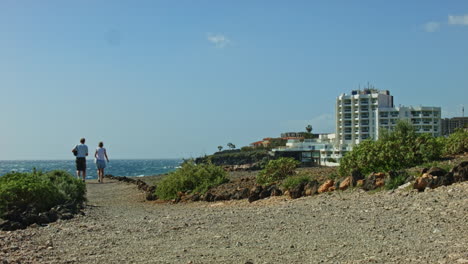 Tenerife-beach-road-tourists-walking-with-a-hotel-and-waves-in-the-background-and-rocky-trail,-road