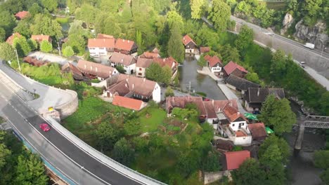 Aerial-shot-of-a-red-car-drives-away-by-a-small-village-surrounded-by-water-and-forest