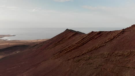 Pull-back-drone-shot-of-a-red-volcano-by-the-ocean