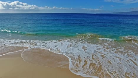 Panning-aerial-shot-in-slow-motion-of-waves-crashing-on-sandy-beach-in-Maui-Hawaii-with-beautiful-sky-and-water