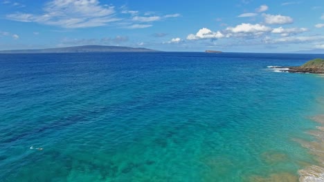 Panning-Luftaufnahme-Eines-Schnorchlers-In-Maui-Hawaii-Im-Wunderschönen-Blauen-Wasser-Mit-Sandstrand-Und-Land-Im-Hintergrund