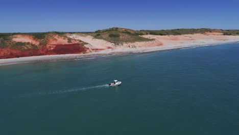 Drone-tracks-a-white-boat-in-far-north-Western-Australia-alongside-red-cliff-tops