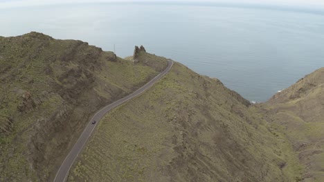 Panorama-drone-shot-of-a-car-driving-through-a-road-in-the-mountains-and-ocean-in-the-background
